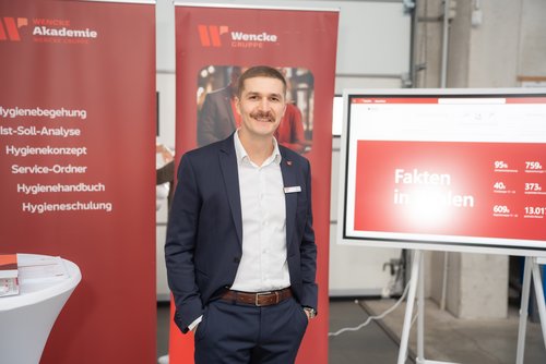 Ein Mann mit braunen Haaren und Schnauzer in blauem Anzug und weißem Hemd, der vor zwei roten Rollups der Wencke Akademie steht